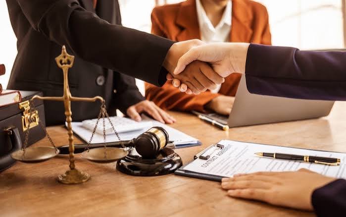 Two attorneys shaking hands across a desk, with scales of justice, a gavel and a signed contract in the foreground
